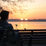 Individual reflecting on dealing with grief while sitting quietly by a serene lake at sunset.